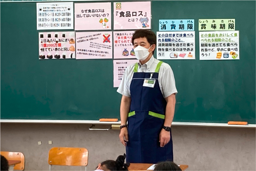 黒板を背景に、男性が授業をしている。黒板には、食品ロスとは何かを書いた文字やイラストのフリップが貼られている。