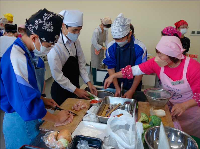 ピンク色のエプロンを着用した食生活改善推進員が、中学生の生徒に調理を教えている。