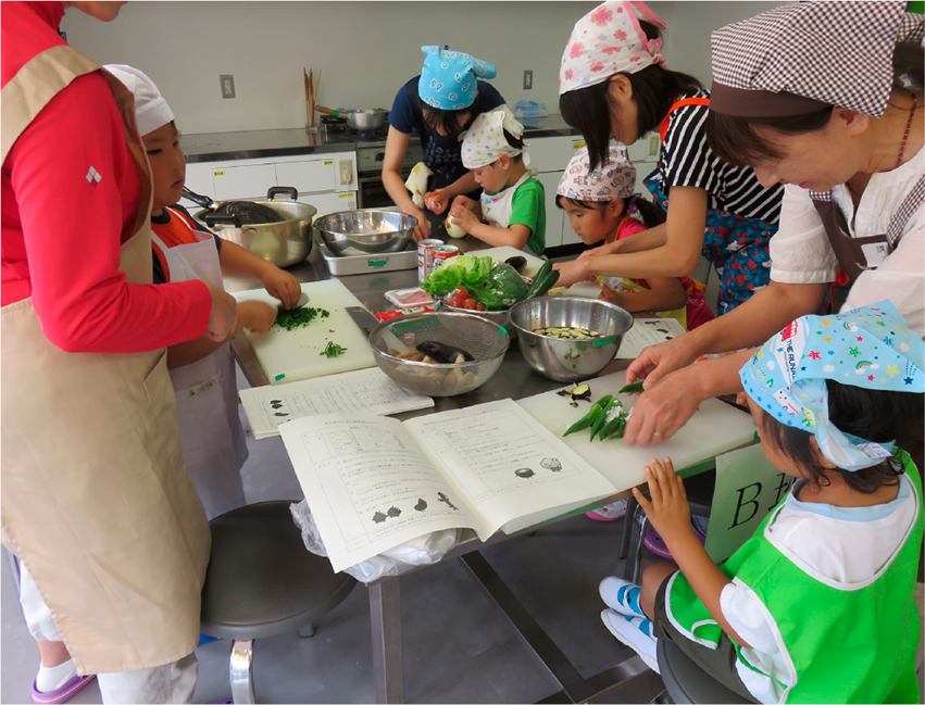 エプロンを着用した親子複数組が調理台で野菜を切ったり、皮をむいたりしている。