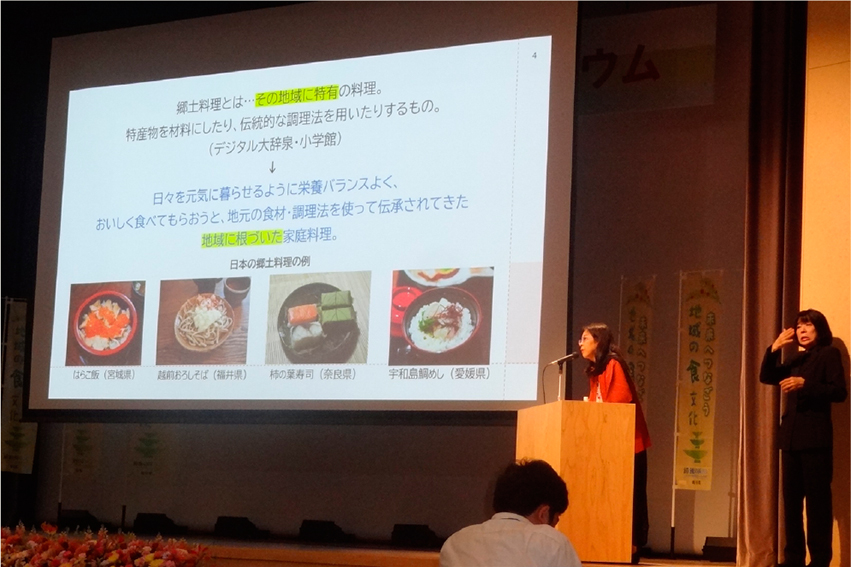 登壇し、話をする女性。大きなスクリーンには郷土料理の写真がみえる。
