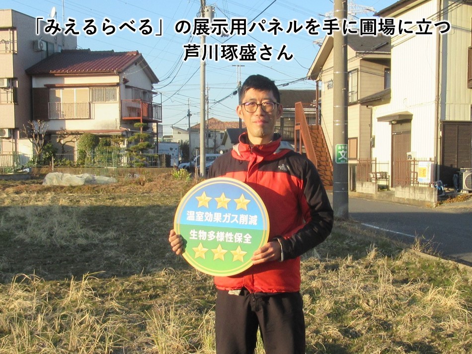 「みえるらべる」の展示用パネルを手に圃場に立つ芦川琢盛さん