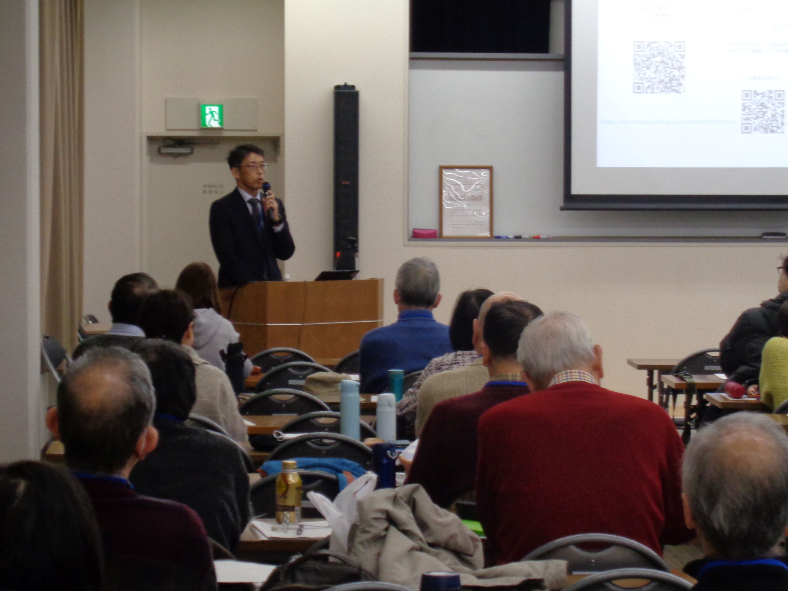 スクリーン横の演台で自己紹介する参事官