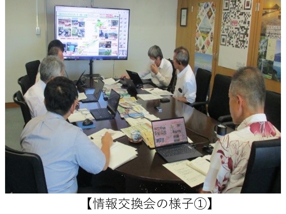 会議室の円卓を囲んで複数の参加者がノートパソコンや資料を手元に置きながら着席し、正面の大型モニターに映し出された資料を見ながら情報交換会を行っている様子。