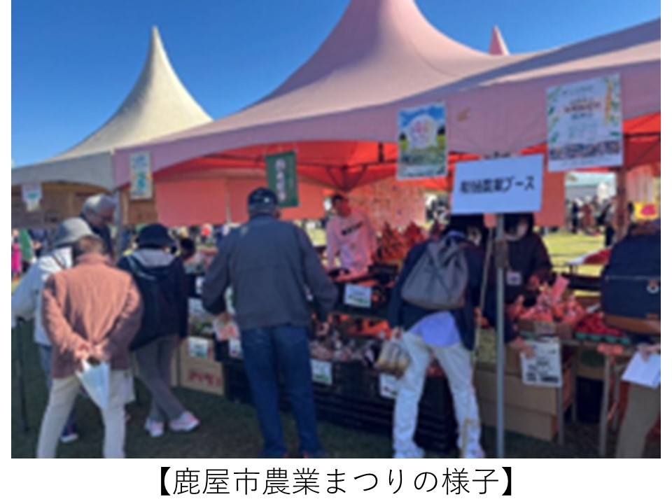 青空の下、白とピンクのテントが立ち並ぶ会場で、「新規農業ブース」と書かれた看板のある販売ブースに多くの来場者が集まり、野菜などの農産物を手に取って見ている鹿屋市農業まつりの様子。