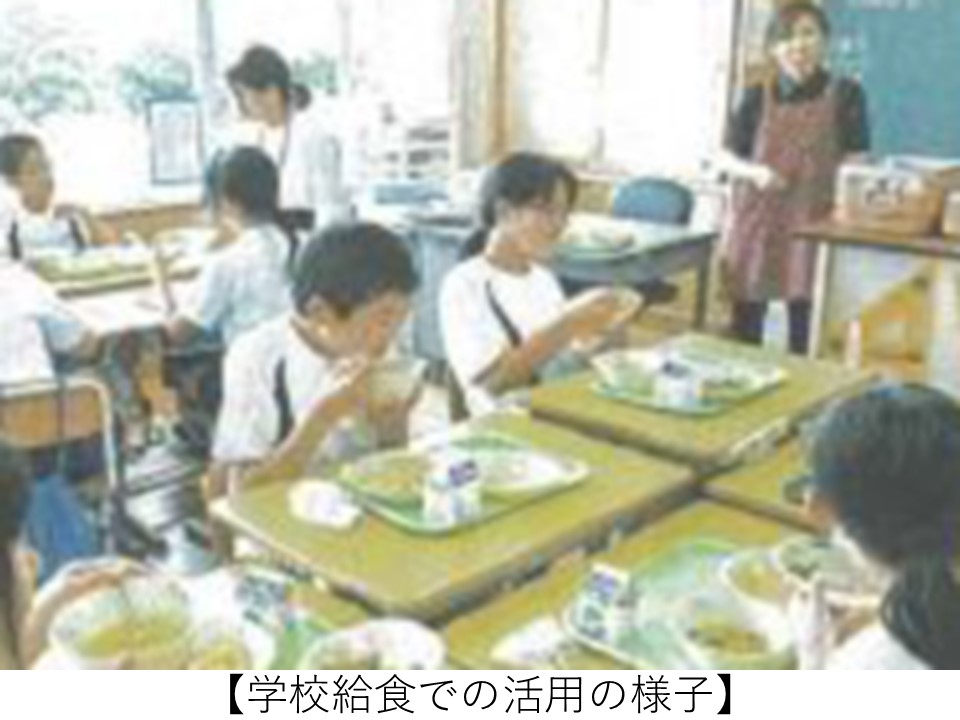 小学校の教室で、児童たちがトレーに乗った給食を食べている様子。エプロン姿の教員が教室前方に立っており、地元農産物を活用した学校給食の実施状況を示している。
