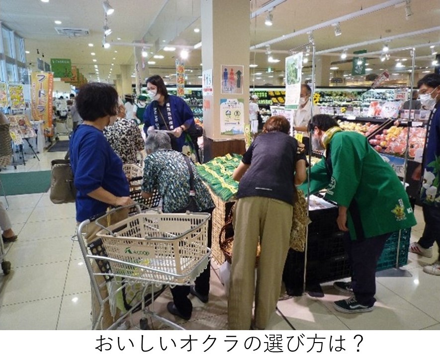 スーパーマーケットの青果売り場で、店員が来店客にIPM栽培オクラの説明をしている様子