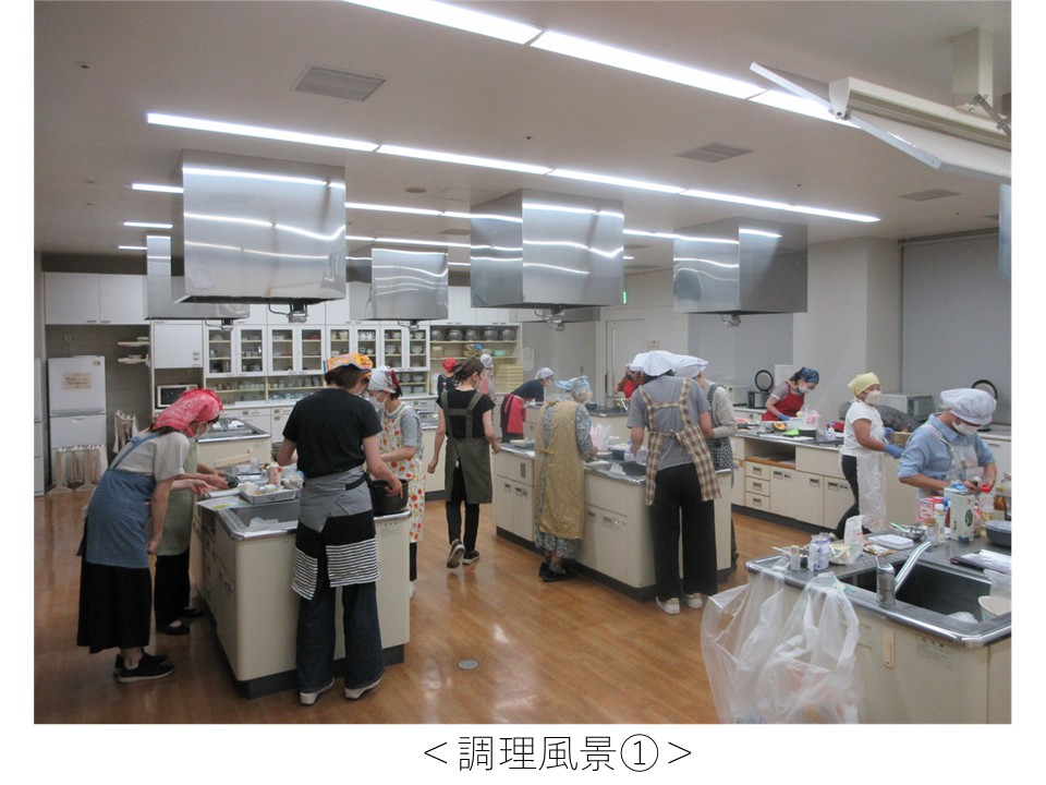 広い調理実習室の中で、エプロンと三角巾・帽子などの衛生帽を着用した10名以上の参加者（主に女性）が、複数の調理台（アイランド型）に分かれて調理作業を行っている。公共施設の調理実習室を使った料理教室で調理実習の場面です。