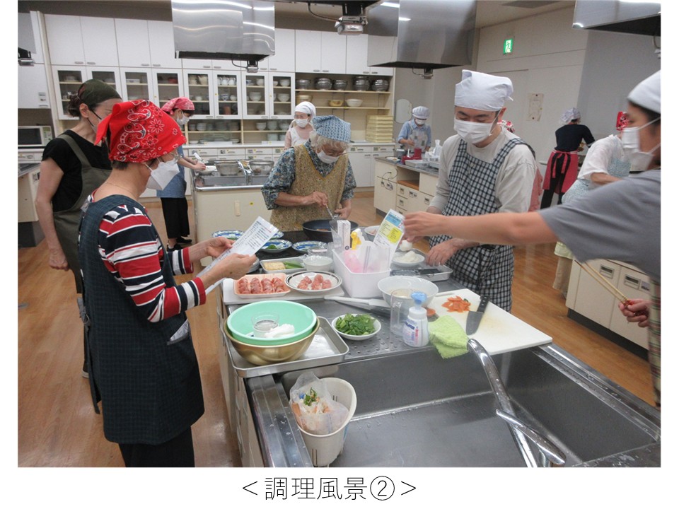 中央の調理台には、肉類・青菜・豆腐などの食材や複数のボウルが並んでいる。白い三角巾と格子柄エプロンを着けた男性が調理台の右側で食材を扱い、向かいでは赤いバンダナと縞模様のシャツ姿の女性がレシピカードを手に確認している。中央付近では花柄エプロンの高齢女性がフライパンを使って調理中。