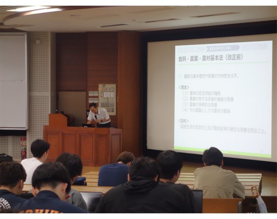 演壇でスライドで説明する職員と聴講する学生