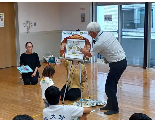 多目的ホールで参加者が床に座り、職員手作りの紙芝居を見ながら説明を聞いています。