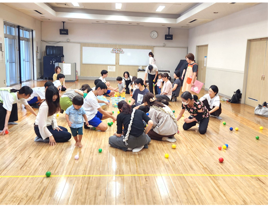 広い多目的ホールの床に大人と子どもが集まり、たくさんのカラーボールを拾ったり並べたりしています。