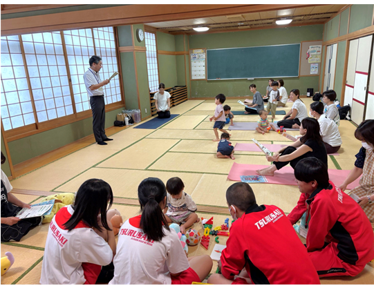 公民館の和室で、前に立った職員が説明をしています。大人は説明を聞き、小さな子どもたちは自由に過ごしています。