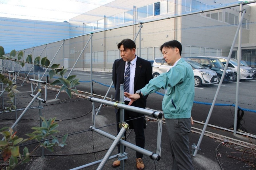 室谷社長(右)から工場敷地内で栽培しているブルーベリーについて説明を受ける中川地方参事官(左)